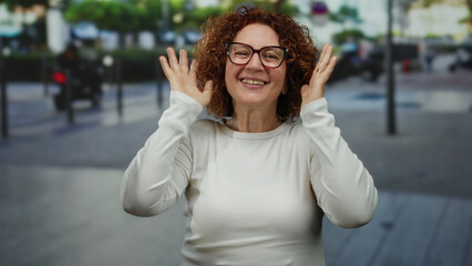 Woman with curly hair wearing glasses in city street covers face in playful moment expressing happiness and surprise with a background of urban scenery blurred behind her.