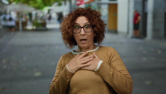 Woman with curly hair and glasses, wearing a brown sweater, expressing surprise on a city street outdoors on a clear day.