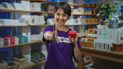 Woman volunteer in purple shirt offers right hand for handshake while holding a red heart stress ball amid shelves of donated goods in a shop building; welcoming kindness.
