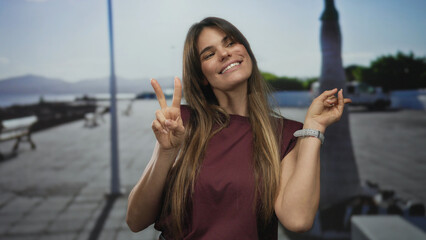 Fototapeta premium Woman smiling on a sunny promenade with sea view, making a peace sign, showcasing vibrant outdoor energy, hispanic cultural essence, and youthful joy.