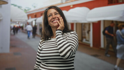 Woman hispanic middle age brunette smiles and covers one eye with her hand while standing on a shopping street with white awnings and blurred pedestrians visible; playful shyness.