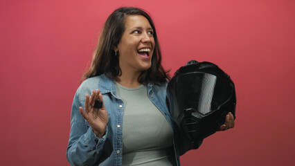 Woman holding a black motorcycle helmet and keys, raised right hand displaying keys, wearing denim jacket, posed against pink studio wall; joy freedom.