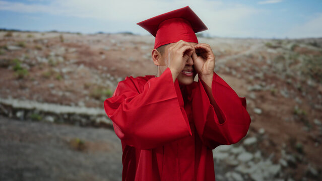 Young man in red graduation gown making binoculars gesture with hands, standing outdoors in a park setting, symbolizing future possibilities and academic achievement.