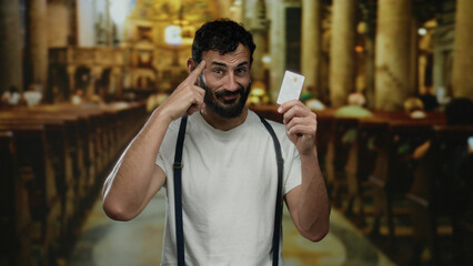 Fototapeta premium Hispanic man with beard showcasing credit card in a catholic church, emphasizing modern financial transactions in a traditional religious setting.