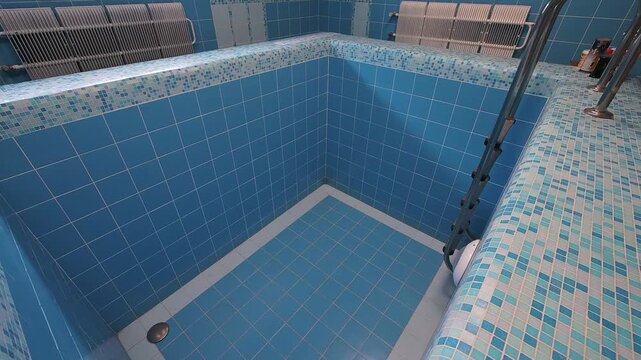 small indoor swimming pool with blue tiled walls and floor, equipped with metal handrails, located under a window with curtains; ceiling lights provide illumination