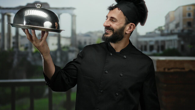 Chef with beard in black uniform holds a silver platter smiling in old town rome with ancient architecture in the background, showcasing culinary artistry.