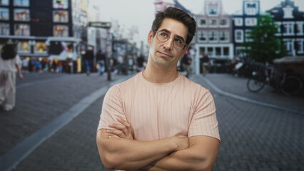 Man with folded arms and visible forearms wearing pale pink t shirt and glasses looking upward on a cobblestone street; thoughtful contemplation.