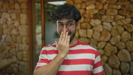 Young man with beard and glasses outdoors in sunny day covers mouth with hand against stone wall in casual striped shirt