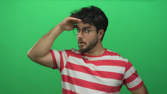 Young hispanic man with beard and glasses mimics looking into the distance with hand on forehead against a vibrant green background wearing a striped shirt.