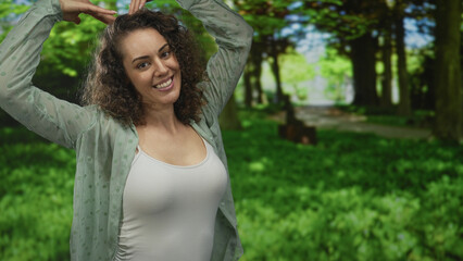 Woman smiling raises arms above head in sunlit forest clearing wearing white tanktop and open shirt; joy.
