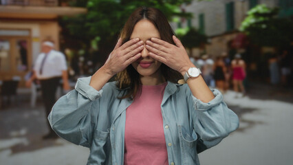 Fototapeta premium Woman surprised on city street wearing denim and pink, covering eyes with playful expression, surrounded by people outdoors, vibrant and lively urban scene, hispanic ethnicity.