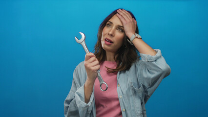 Woman expressing surprise holding wrench against vibrant blue background depicts hispanic young female in a casual shirt using a tool, emphasizing intrigue and emotion.