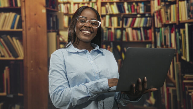 Woman wearing glasses holding laptop and typing while smiling among bookshelves in a library building; confidence learning concentration.
