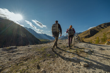 Fototapeta premium Two hikers with backpacks walk an autumn mountain road overlooking a valley with high peaks and colorful forest. Rear view captures their shadows on the path under a cloudy sky.