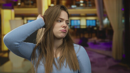 Woman touching hair with hand while tilting head near a yellow lamp in building hallway with pursed...