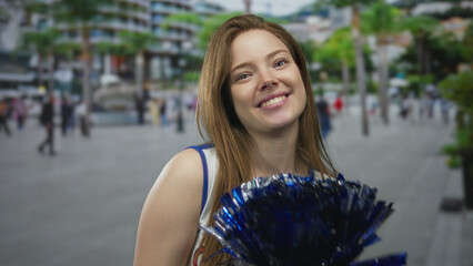 Young woman cheerleader waves metallic blue pompom on city street with blurred urban buildings;...