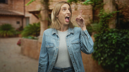 Obraz premium Woman in denim jacket with arms crossed smiling and looking up on a stone street courtyard near a rustic wall; curiosity exploration.