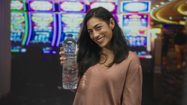 Woman holding a water bottle with visible hand and smile near slot machines in a building casino gaming floor; satisfaction refreshment.