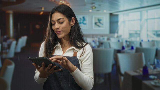 Woman holding tablet and flashing peace sign in restaurant dining room while wearing apron and smiling, tapping screen; service hospitality joy.