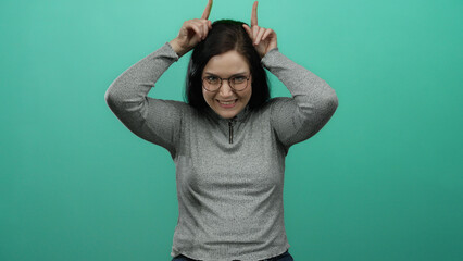 Brunette woman making horn gesture with fingers against a vibrant green background while smiling playfully in casual attire.