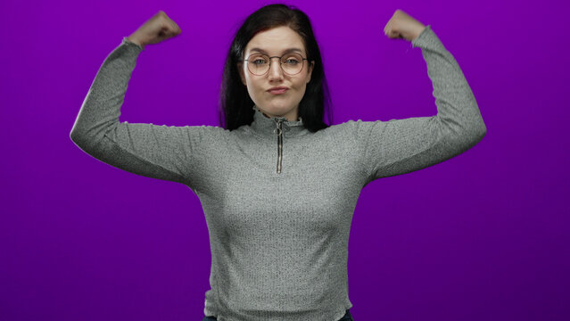 Confident woman in glasses posing with raised fists against a vibrant purple background, wearing a grey sweater, showcasing empowerment, strength, and determination.