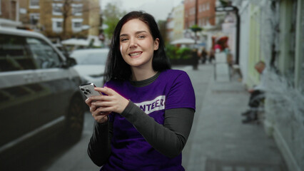 Fototapeta premium Woman with brunette hair smiling while holding a smartphone on a street in a volunteer shirt outdoors, surrounded by blurred vehicles and buildings in an urban area.