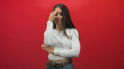 Young woman covers mouth with hand and crosses arm against a red studio backdrop; surprise...