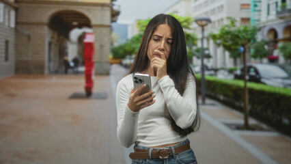 Young hispanic woman reading smartphone with furrowed brow and hand to chin on street near red sculpture and archway; concern message.