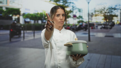 Woman chef in white coat holds green cooking pot with wooden handles and points finger forward on a busy street, steady gesture and direct gaze; focus determination.