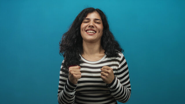 Woman in striped shirt clenching fists with head tilted back and exposed neck in a teal studio; excitement.