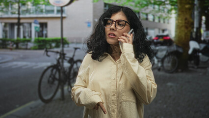 Woman holding smartphone, pointing finger to screen, wearing glasses and beige shirt on a street with parked bicycles and cars; urgent communication concern.