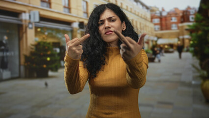 Fototapeta premium Young woman shows both middle fingers to camera on a city street lined with shops and pedestrians; defiance.