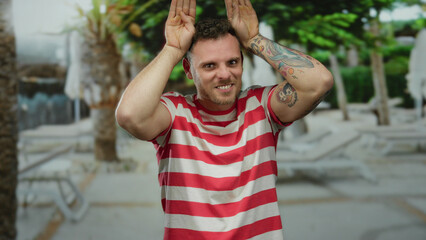 Happy man with tattoos and striped shirt playfully posing outdoors in an urban area with blurred trees in the background on a sunny day.