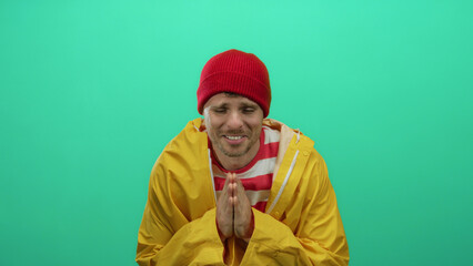Young man wearing a red beanie and yellow jacket poses against a vibrant green background, exuding an attractive and playful vibe with his charming expression.
