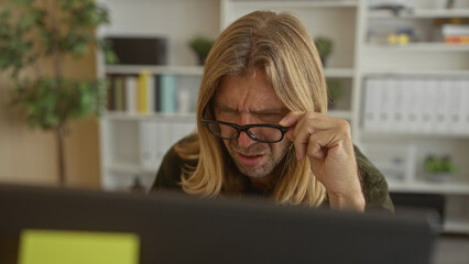 Blond man with long hair adjusts glasses frowns and leans forward toward a computer screen in a building; confusion.