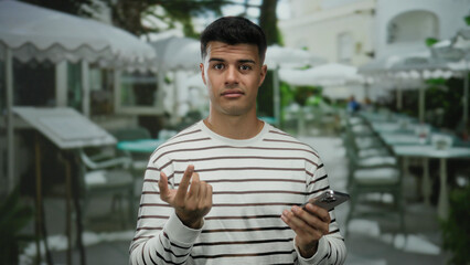Young hispanic man on terrace holding phone looks contemplative outside a city cafe wearing striped shirt surrounded by tables and chairs in an urban outdoor setting