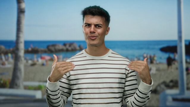 Handsome young man gesturing in a striped shirt at a vibrant seaside beach with palm trees and ocean in the background, conveying a casual summer vibe outdoors.