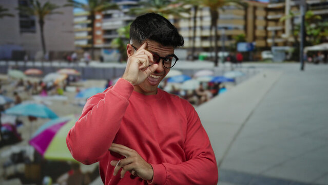 Young man in glasses smiling at seaside with vibrant beach background and palm trees, wearing a red sweater, embodying cheerful vibe outdoors.