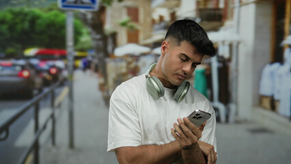Young man with headphones checking smartphone on bustling city street during daytime.