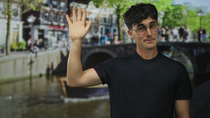 Man waves hand with open palm on street in amsterdam by canal and bridge, wearing glasses and black shirt; friendly greeting.