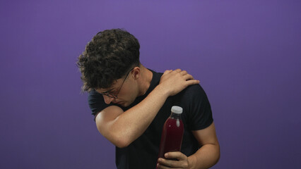 Young hispanic man wearing glasses and black t shirt holding a red bottle of juice and touching shoulder in purple studio; discomfort recovery.
