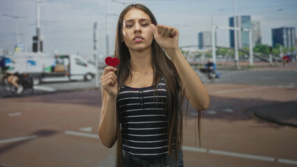 Fototapeta premium Teen woman holds a small red heart in one hand while raising a closed fist toward the camera on a city street; heartbreak longing.