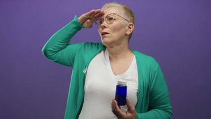 Woman holding blue pill bottle with hand to forehead shielding eyes in purple studio; health concern seeking relief.