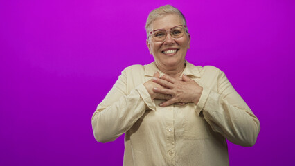 Middle aged woman with hands over heart smiling and reaching forward in magenta studio; tender warmth gratitude connection.