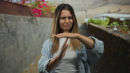 Woman with visible hands forming a time out hand gesture on a street beside a stone wall and planter; disgust.