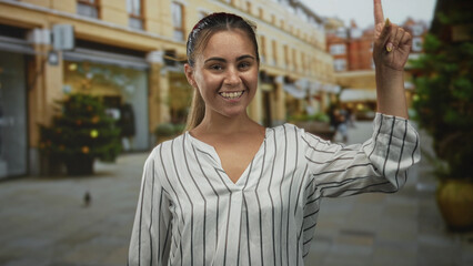 Hispanic woman pointing finger upward on street near shops, smiling with open mouth and visible hand gesture; joy celebration.