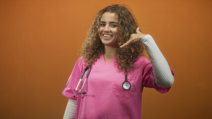 Woman doctor in pink scrubs with stethoscope making phone gesture with right hand in studio; friendly approachable care.
