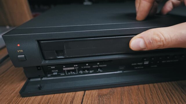 Male hand inserts black VHS cassette into motorized slot of vintage VCR player. Finger presses control button on lower panel amidst wooden shelf background.