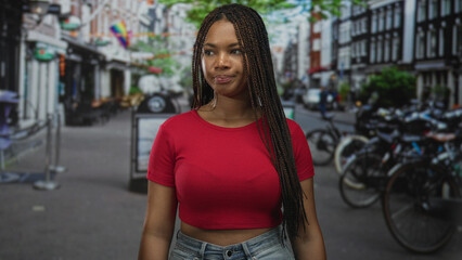 Naklejka premium Woman with braided hair and visible breasts standing on a city street in a red cropped shirt; quiet confidence.