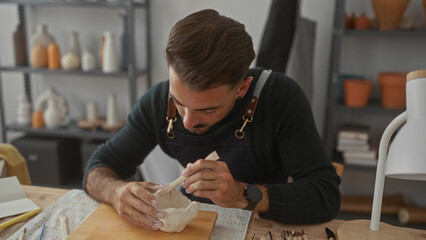 Hispanic artisan man carving clay bowl with wooden modeling tool in a pottery studio workshop, shelves of ceramics behind him; creativity.
